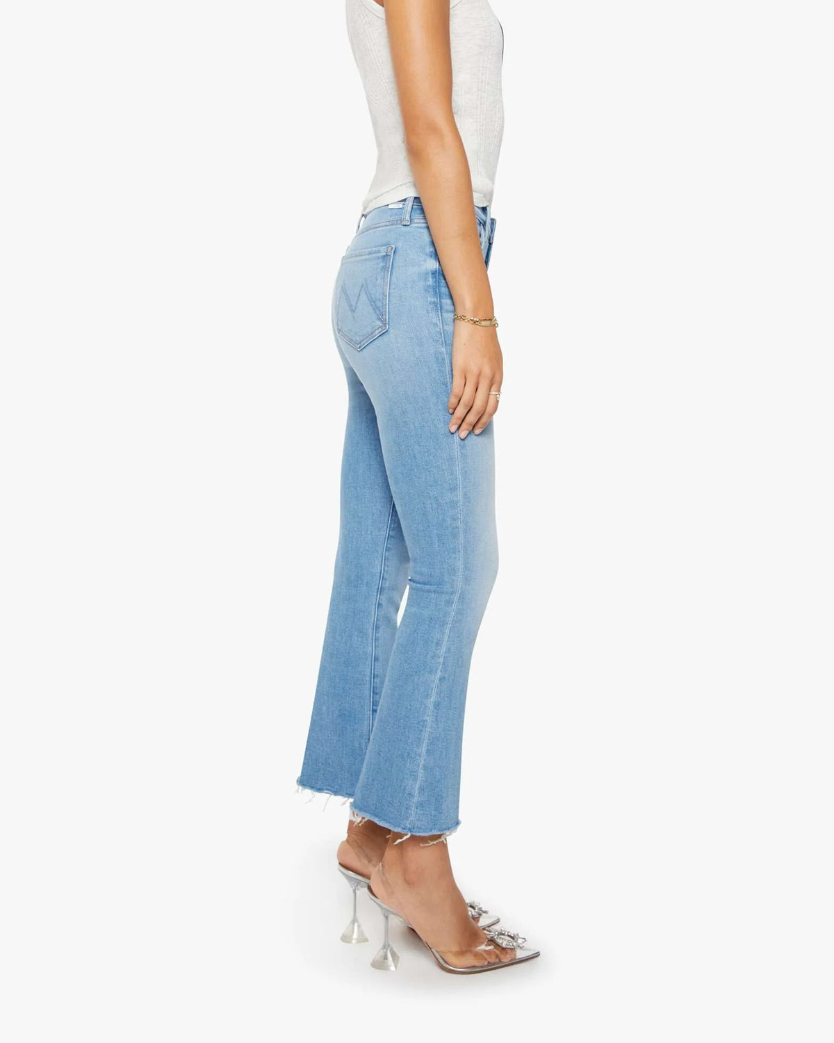 A woman in profile wears Mother’s The Outsider Flood Fray light blue mid rise cropped flare jeans with raw hems, a white sleeveless top, and unique clear high-heeled sandals against a white background.
