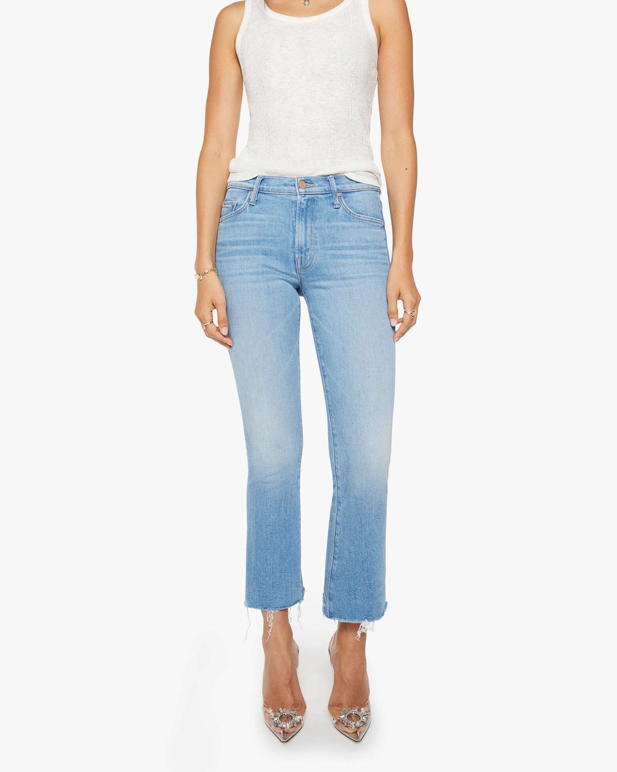 A person wears Mother’s The Outsider Flood Fray light blue straight-leg jeans with frayed hems, a sleeveless white top, and pointed-toe clear heels, standing against a white background.