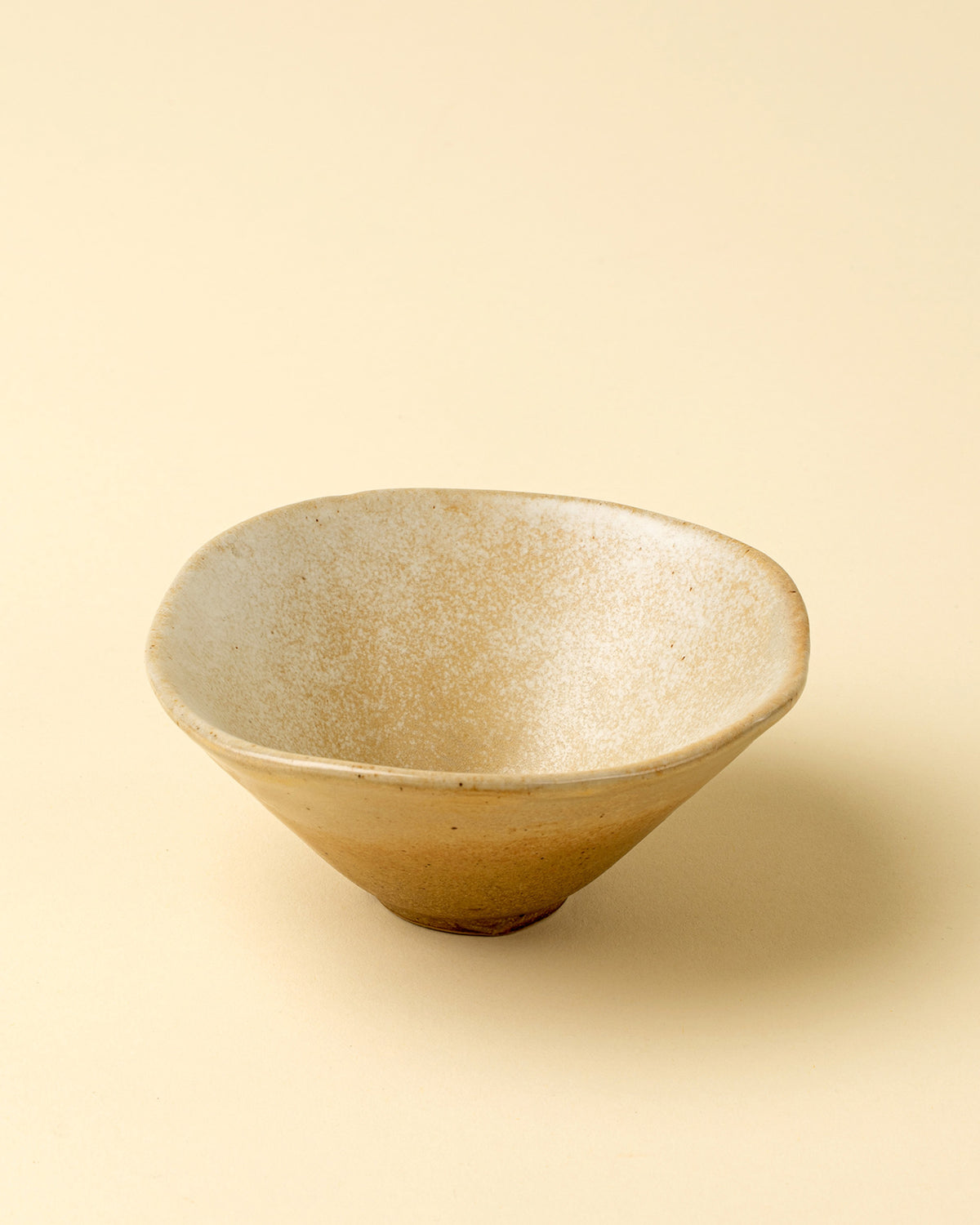 The Indaba 4-6490 Petra Porcelain Bowl is a handmade, shallow tan ceramic bowl with a slightly uneven rim, smooth matte finish, and subtle speckled patterns, shown on a light beige background for an elegant touch.