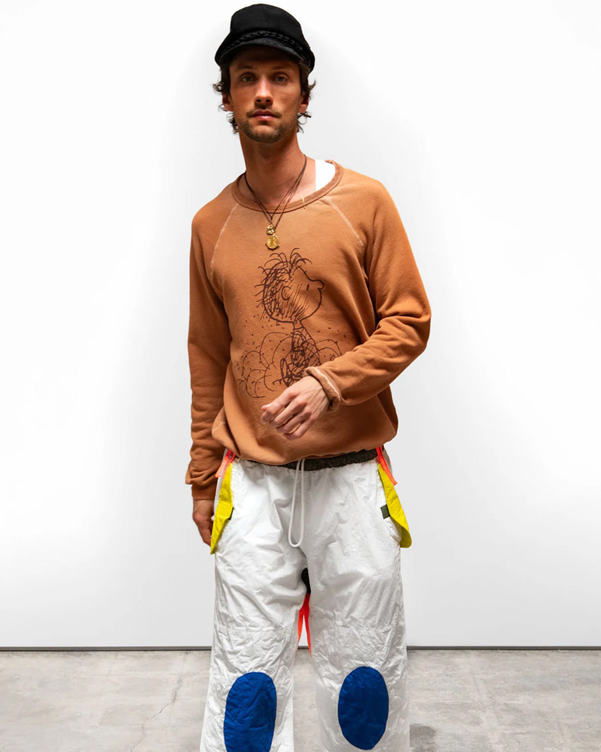 A man in a black cap, white pants with blue knee patches and yellow accents, and a Free City (sparrow, LLC) BasicGoodness Schulz Raglan sweatshirt stands against a plain white background.