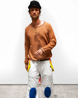 A man in a black cap, white pants with blue knee patches and yellow accents, and a Free City (sparrow, LLC) BasicGoodness Schulz Raglan sweatshirt stands against a plain white background.