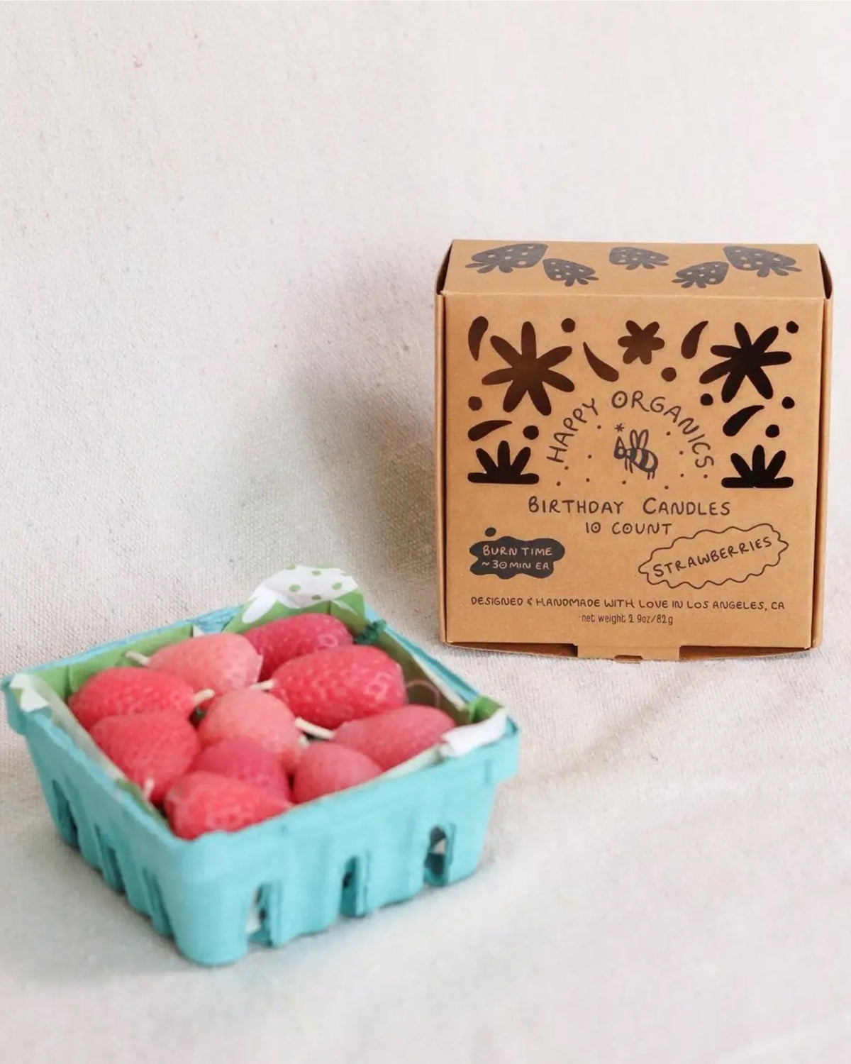 A blue carton contains Faire Pink Strawberry Birthday Candles, shaped like strawberries, beside a brown box labeled "Faire Pink Strawberry Birthday Candles 10 Count," featuring hand-drawn designs and eco-friendly candle info.