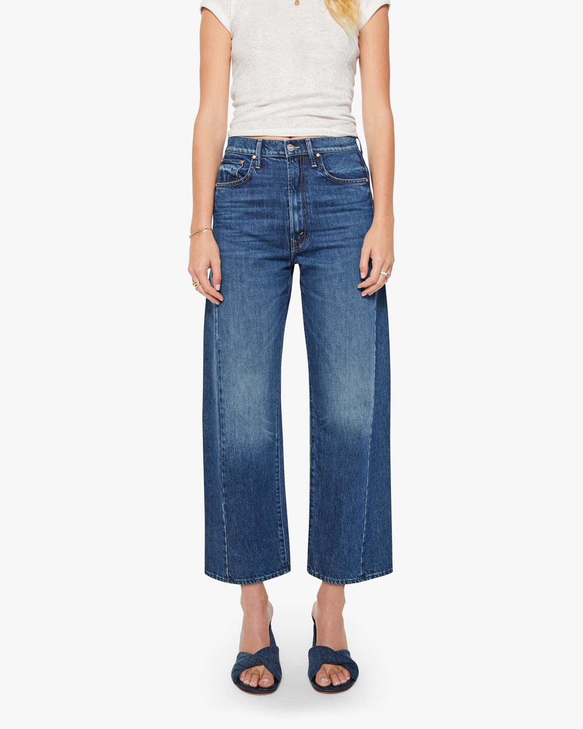 A person wearing a white short-sleeve top, Mother’s The Half-Pipe Ankle high-waisted jean in SUPERIOR denim, and dark blue sandals stands against a plain white background.