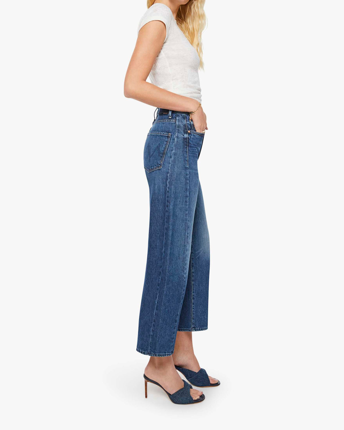 A woman stands in profile against a plain white background, wearing The Half-Pipe Ankle high-waisted jeans in a dark blue wash by Mother, paired with a short-sleeve white top and navy heeled sandals.