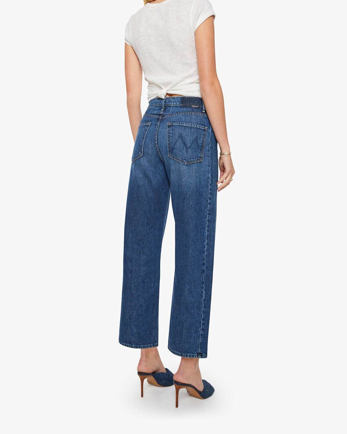 A woman stands with her back turned, wearing a white short-sleeve top, blue heeled mules, and Mother’s The Half-Pipe Ankle high-waisted jeans in a dark blue wash with wide legs.