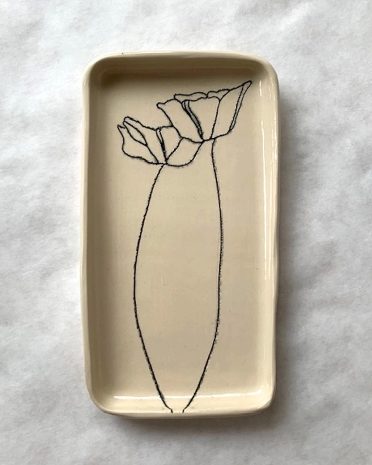 The Floral Small Tray by Faire is a rectangular cream ceramic tray featuring a minimalist black line drawing of two poppy flowers, one large and one small, etched by hand. It is displayed on a white textured surface.