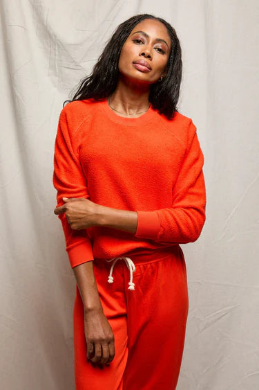 A woman with long dark hair wears the Perfectwhitetee Ziggy Reverse LS Sweatshirt and matching pants in bright orange, standing before a light backdrop with one arm across her body.