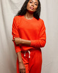 A woman with long dark hair wears the Perfectwhitetee Ziggy Reverse LS Sweatshirt and matching pants in bright orange, standing before a light backdrop with one arm across her body.