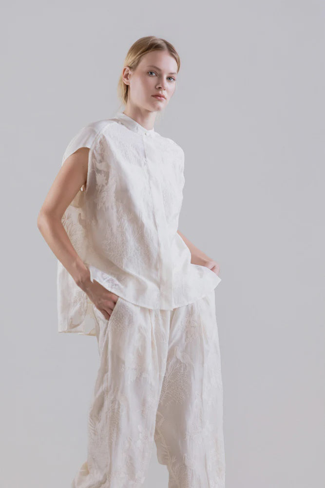 A person stands against a light gray background, wearing the Psophia 2705 Womens Top—a loose white embroidered Tencel blouse with flowing sleeves—paired with matching wide-legged pants, one hand casually in a pocket.