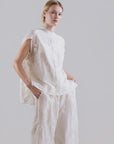 A person stands against a light gray background, wearing the Psophia 2705 Womens Top—a loose white embroidered Tencel blouse with flowing sleeves—paired with matching wide-legged pants, one hand casually in a pocket.