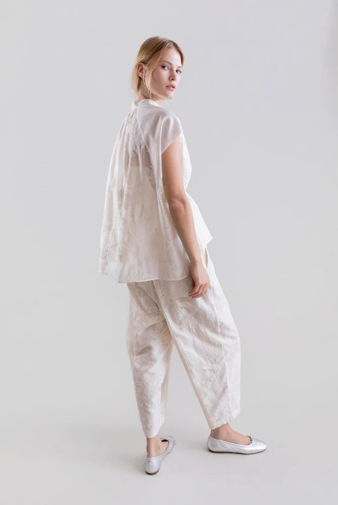 A woman stands against a plain light background, wearing the Psophia 2705 Womens Top—an embroidered Tencel blouse with flowing sleeves—and matching textured white pants. She looks over her shoulder and pairs the outfit with white flats.