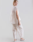 A woman stands against a plain light background, wearing the Psophia 2705 Womens Top—an embroidered Tencel blouse with flowing sleeves—and matching textured white pants. She looks over her shoulder and pairs the outfit with white flats.