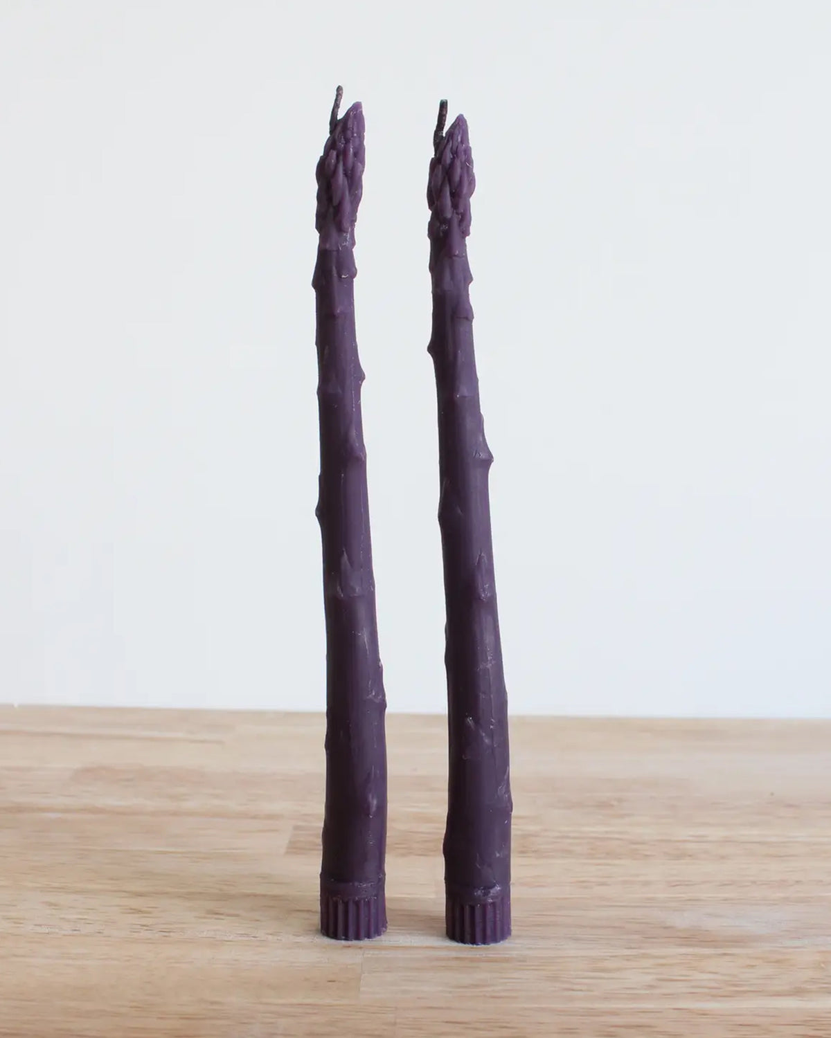 Two Faire Purple Asparagus Candle Tapers stand upright on a light wooden surface against a plain off-white background.