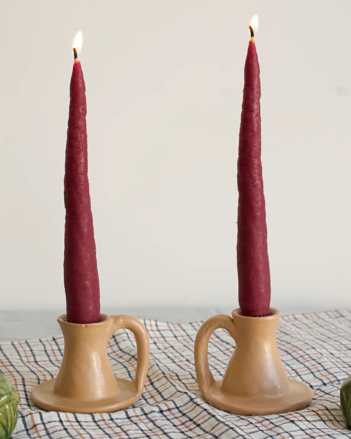 Two Purple Carrot Candle Tapers by Faire are lit and set in tan ceramic holders with handles, resting on a checkered cloth against a plain, light background.