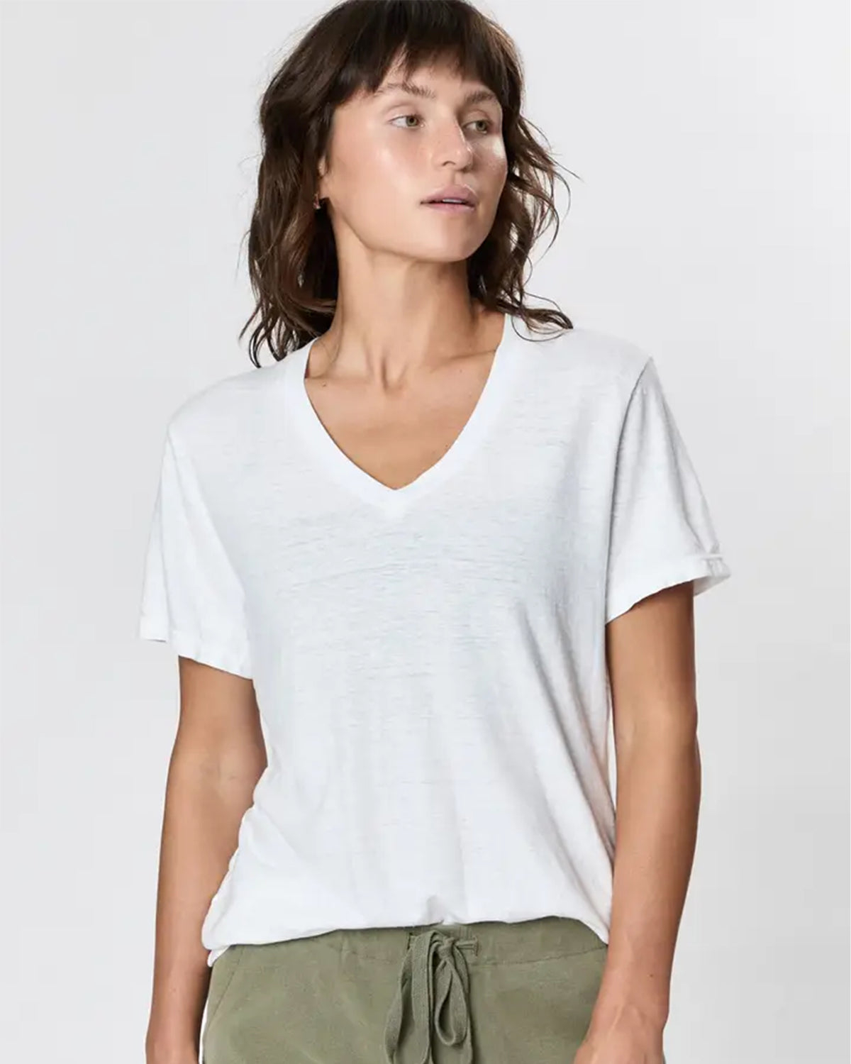 A person with wavy brown hair wears the Faire Hemp V Neck, a loose white T-shirt, and olive green drawstring pants, standing against a plain light background and looking slightly to the side.