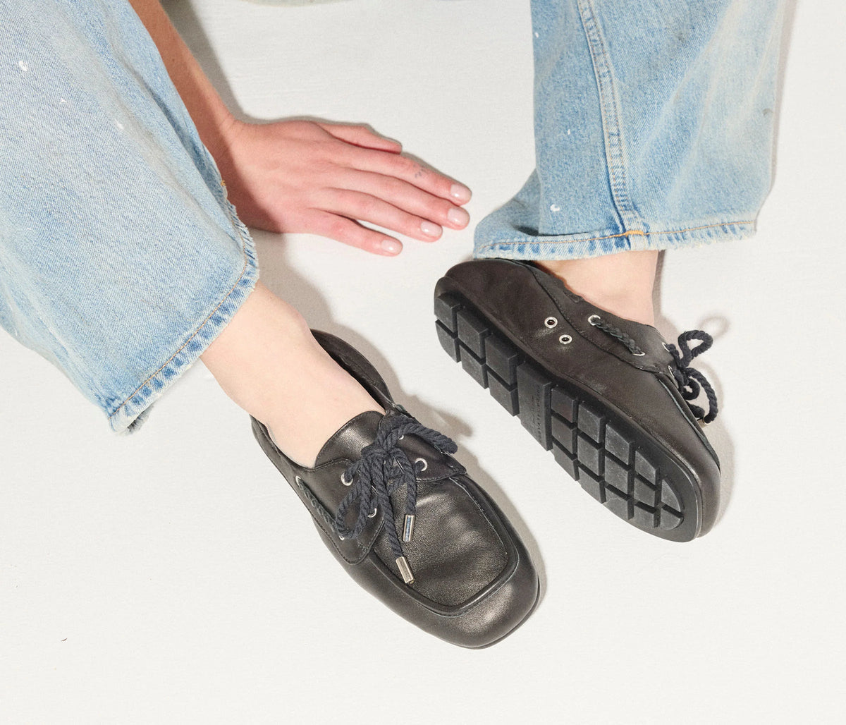 A person wearing light blue jeans and Freda Salvador RIA Driver Loafers in black Nappa leather with thick soles sits on a light surface, legs extended, hands resting on the floor.