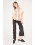 A woman with light brown hair poses casually against a white background, wearing a cream embroidered short-sleeve shirt, black pants with gold buttons, and Freda Salvador’s RIA Driver Loafers in stylish black Nappa leather.