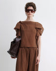 A person wears the Rachel Comey Rivera Top in brown crinkled fabric with flutter sleeves, matching brown pants, and sunglasses, holding a large dark brown bag against a plain white background.