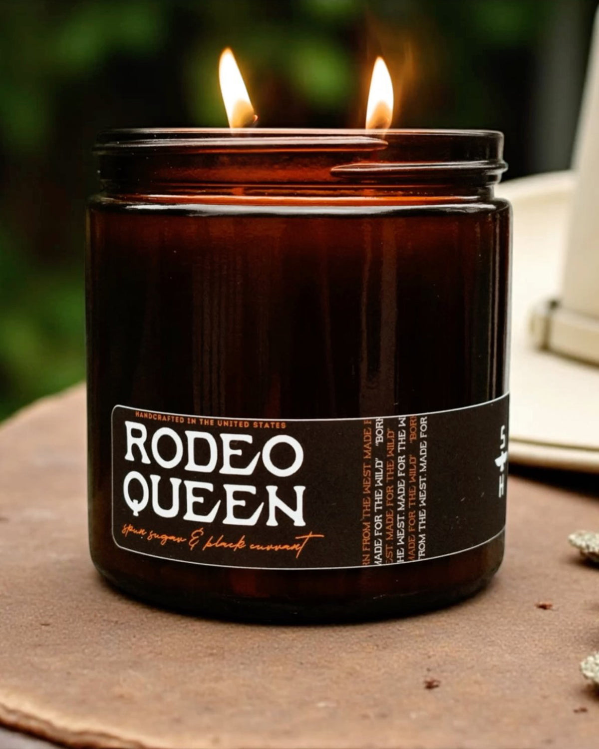 Brown jar Rodeo Queen Candle with twin flames by Faire, soy coconut wax, glowing outdoors on a round surface.