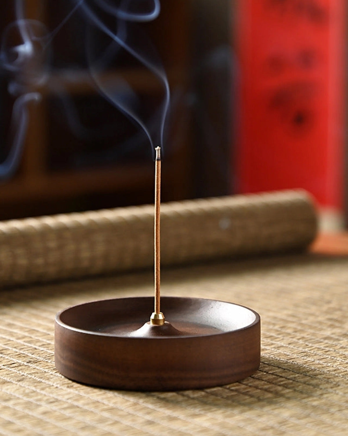 The Faire Walnut Wood Incense Burner emits a thin trail of smoke, placed beside handcrafted ceramic plates featuring traditional Chinese designs. The setup sits on a woven mat with blurred bamboo and red panel backgrounds.