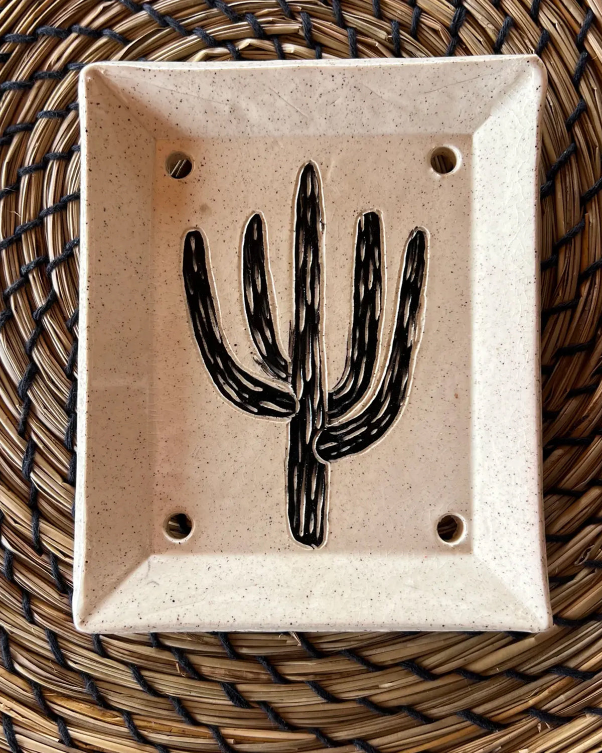 The Saguaro Soap Dish is a handmade sgraffito piece with a black cactus design at its center and four drainage holes, presented on a woven circular mat featuring brown and black patterns.
