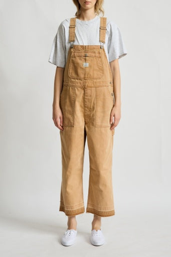 A model showcases the Denimist RELAXED OVERALL—a tan, 100% cotton piece featuring adjustable buckle straps—layered over a loose gray T-shirt and paired with white sneakers. Against a plain white background, the overall’s relaxed-leg silhouette highlights its laid-back style as the model stands with arms comfortably at their sides and face partially out of frame.