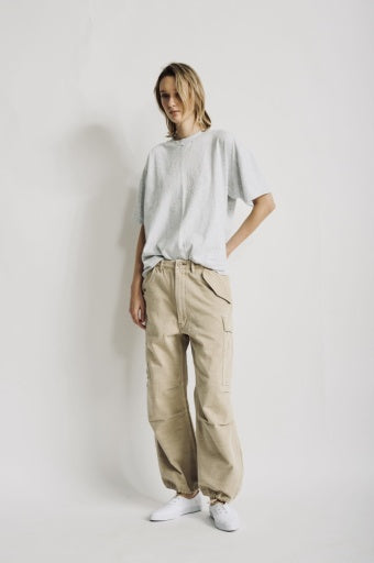 A person stands against a plain white background wearing a relaxed-fit light gray t-shirt, Denimist CROPPED CARGO beige utility pocket pants, and white shoes, with one hand in their pocket.