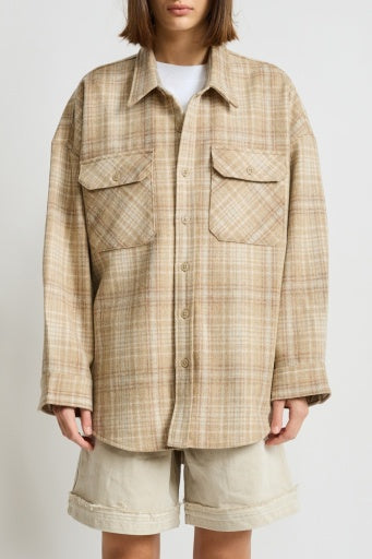 A person wears an oversized beige plaid Denimist RORY SHIRT JACKET with flap pockets over a white top and beige knee-length shorts, standing against a plain light background.