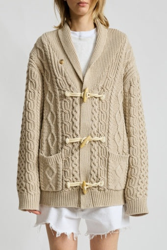 A person wears the Denimist TARYN TOGGLE SWEATER—an oversized, beige cable-knit with front pockets and slip buttons—over a white tee and frayed hem shorts, standing against a plain background.