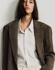 A person with straight, shoulder-length dark brown hair wears the Denimist HYANNIS DECONSTRUCTED BLAZER over a white collared shirt and cream pants, standing against a plain light background.