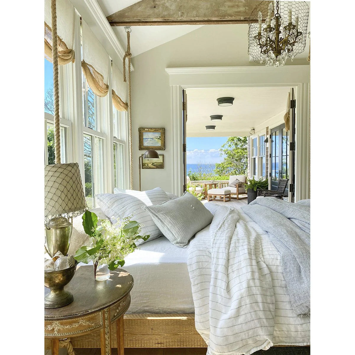 The image presents a well-lit bedroom showcasing white bedding from the Lake Linen Collection, accompanied by a bedside table featuring a lamp and vase. Through large windows, you can see views of a porch with seating arrangements and the serene blue ocean in the distance, framed by lush greenery. A chandelier hangs gracefully from the ceiling, enhancing the room's elegance with its presence next to POM POM's Blake Big Pillow 28x36.