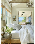 The image presents a well-lit bedroom showcasing white bedding from the Lake Linen Collection, accompanied by a bedside table featuring a lamp and vase. Through large windows, you can see views of a porch with seating arrangements and the serene blue ocean in the distance, framed by lush greenery. A chandelier hangs gracefully from the ceiling, enhancing the room's elegance with its presence next to POM POM's Blake Big Pillow 28x36.
