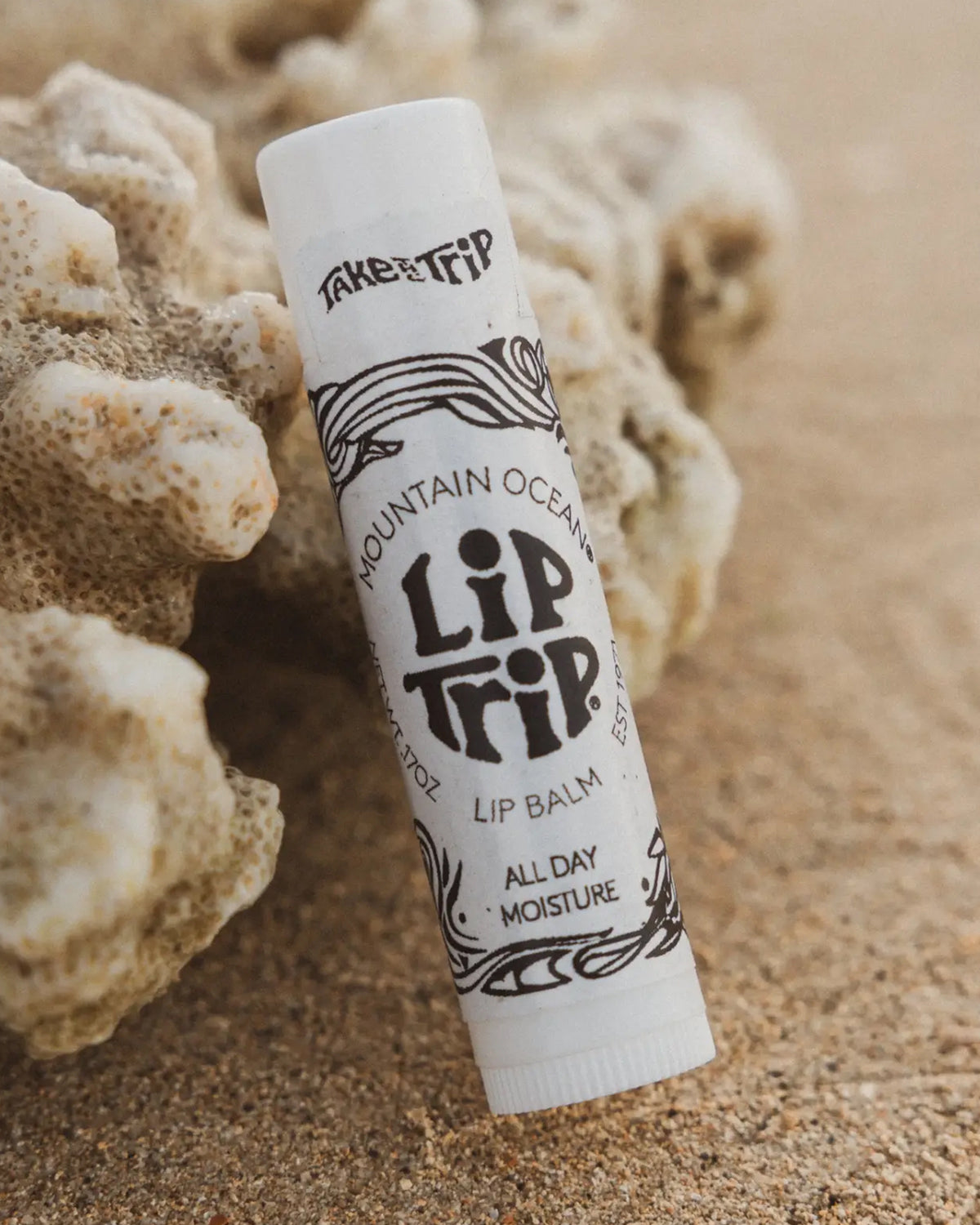 A tube of Faire Skin Trip Lip Balm sits on sandy ground by coral. The label reads "Faire Skin Trip Lip Balm All Day Moisture" with black decorative designs on a white background.