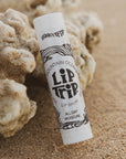 A tube of Faire Skin Trip Lip Balm sits on sandy ground by coral. The label reads "Faire Skin Trip Lip Balm All Day Moisture" with black decorative designs on a white background.