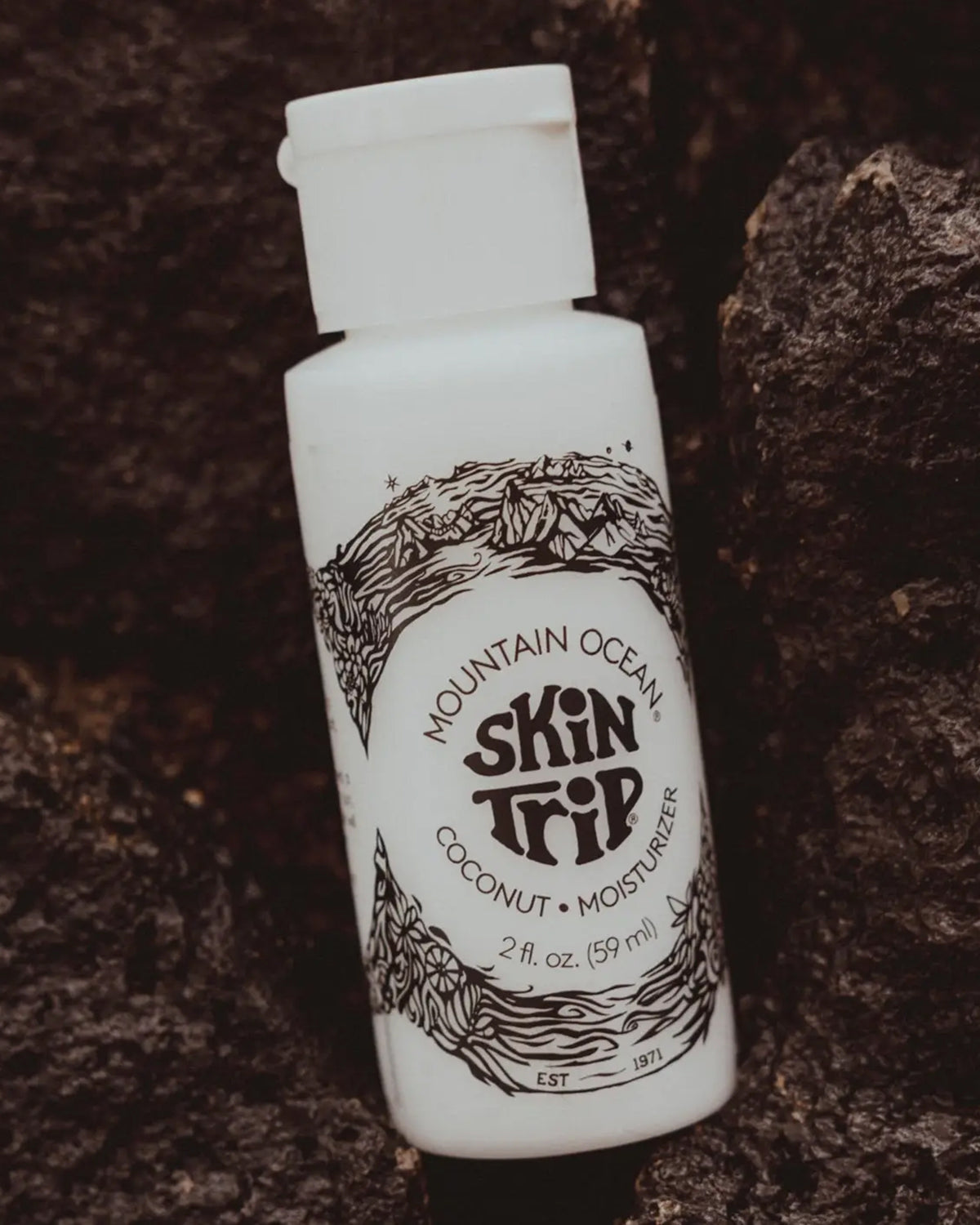 A white bottle of Faire Skin Trip Travel Size sits on dark, textured rocks. This coconut-scented lotion features black mountain and wave art, offering natural skincare in a convenient travel-size moisturizer.