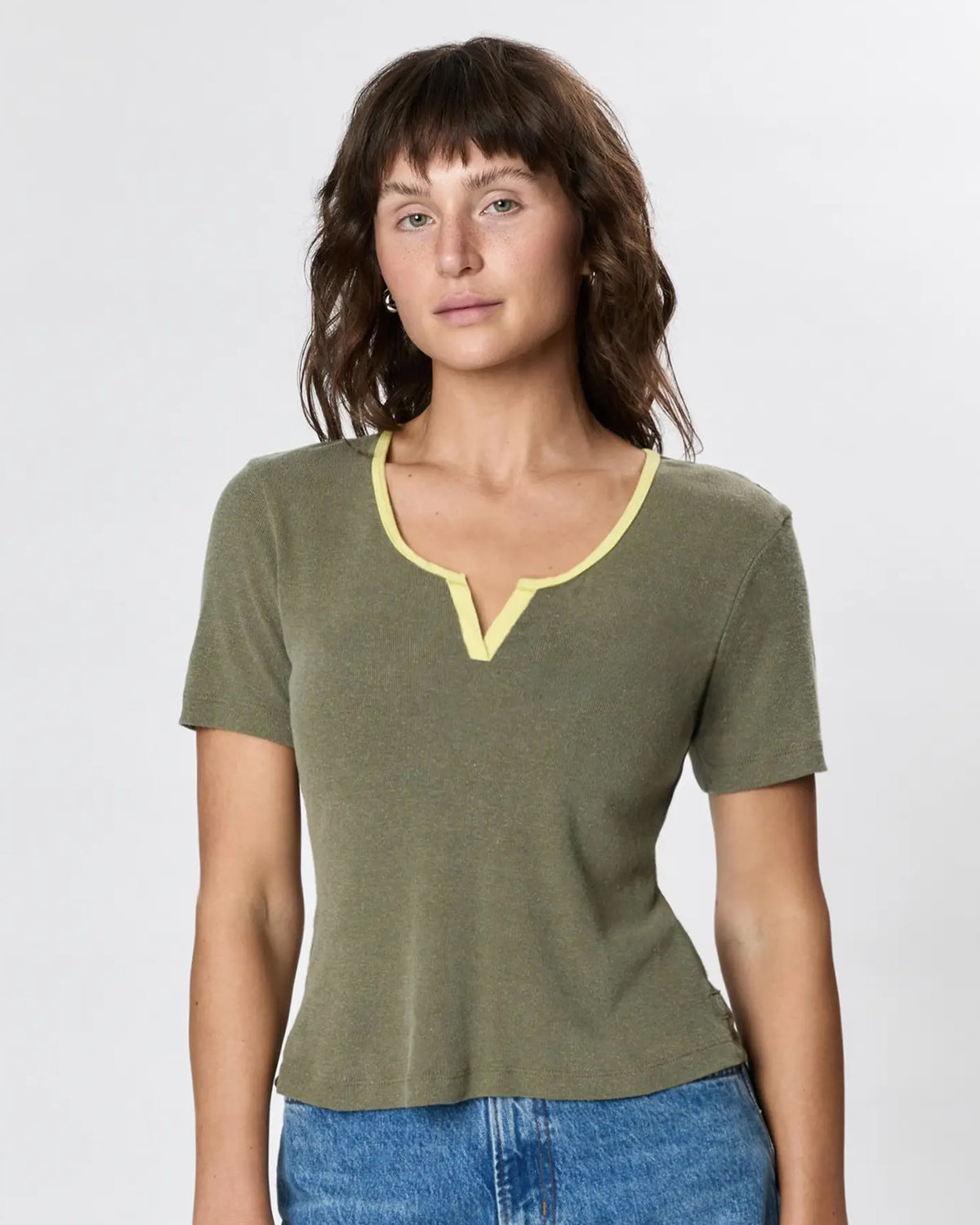 A woman with wavy brown hair wears Faire's Stardust Hemp Top—an olive green organic cotton short-sleeve shirt with a light yellow-trimmed notch neckline—paired with blue jeans, standing against a plain light background.
