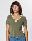 A woman with wavy brown hair wears Faire's Stardust Hemp Top—an olive green organic cotton short-sleeve shirt with a light yellow-trimmed notch neckline—paired with blue jeans, standing against a plain light background.