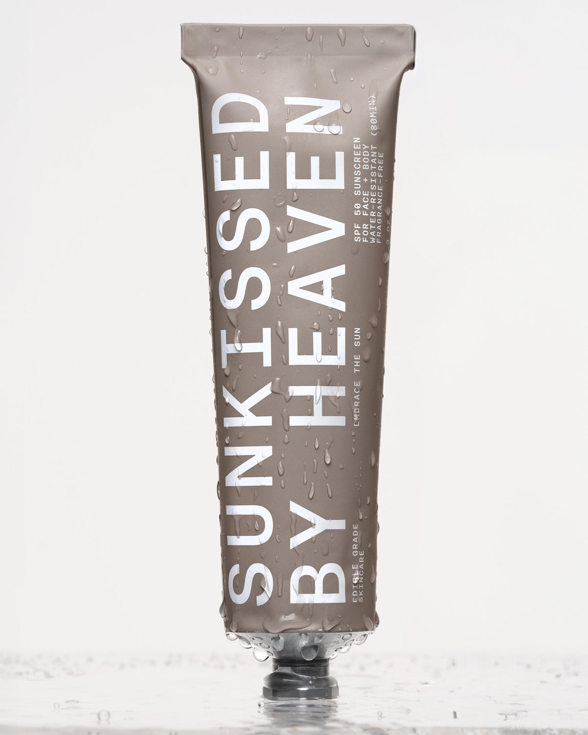 A taupe-colored tube of Faire Tallow Sunscreen SPF 50 stands upright with water droplets on its surface, offering broad-spectrum protection for sensitive skin against a light, reflective background.