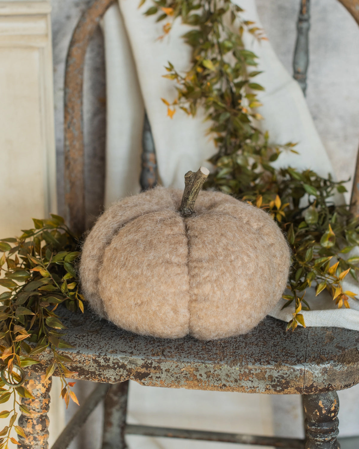 Tan fabric pumpkin with twig stem by Faire on rustic chair, surrounded by leafy garland; cozy fall accent.