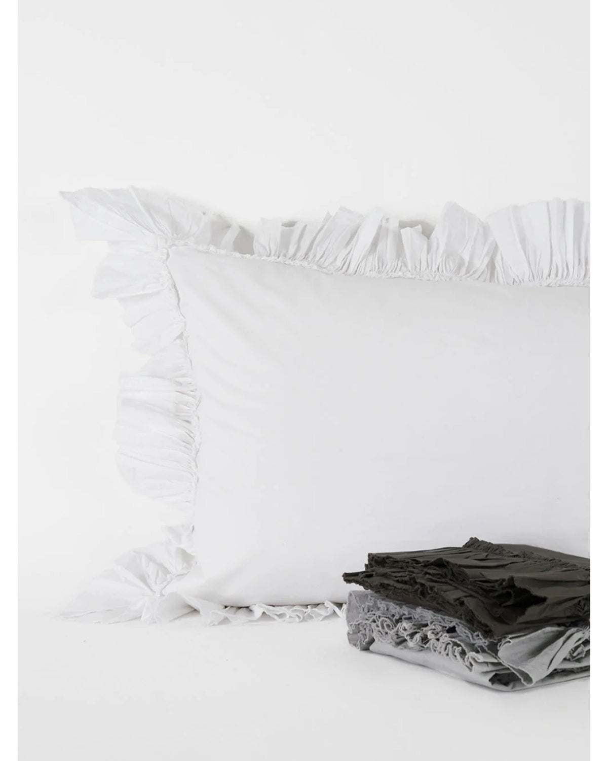 The Tat Cotton Sham Euro by Matteo, in white cotton with ruffled edges, is displayed against a white background. In front, gray and black fabric items highlight the soft percale weave and 225 thread count.