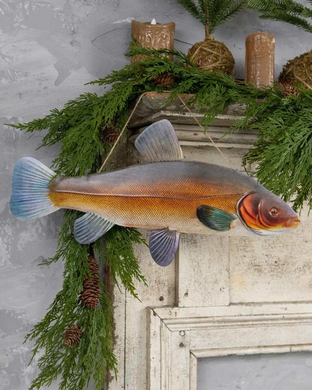18.25″ TENCH FISH wall decor by Faire displayed above a rustic mantel with greenery, pine cones, and candles.
