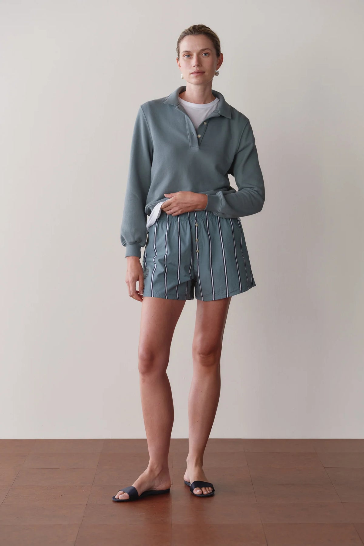 A person stands indoors against a plain light wall and wooden floor, wearing Donni's The Stripe Pop Boxer in blue-and-white stripes, paired with a light blue pullover, white t-shirt, and dark slide sandals.