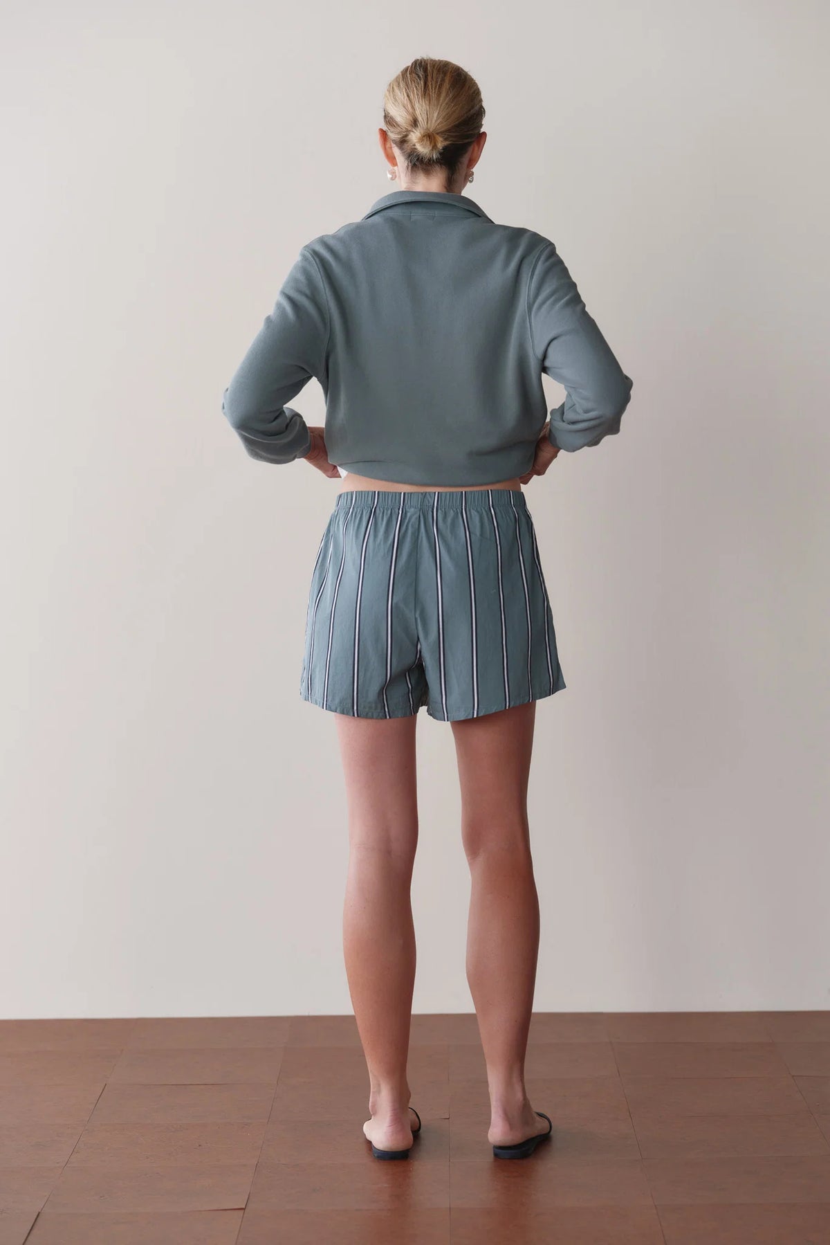 A person stands facing a blank wall, wearing a cropped, long-sleeve gray top, Donni's The Stripe Pop Boxer in blue and white stripes, and black sandals. Their hair is pulled back in a low bun.