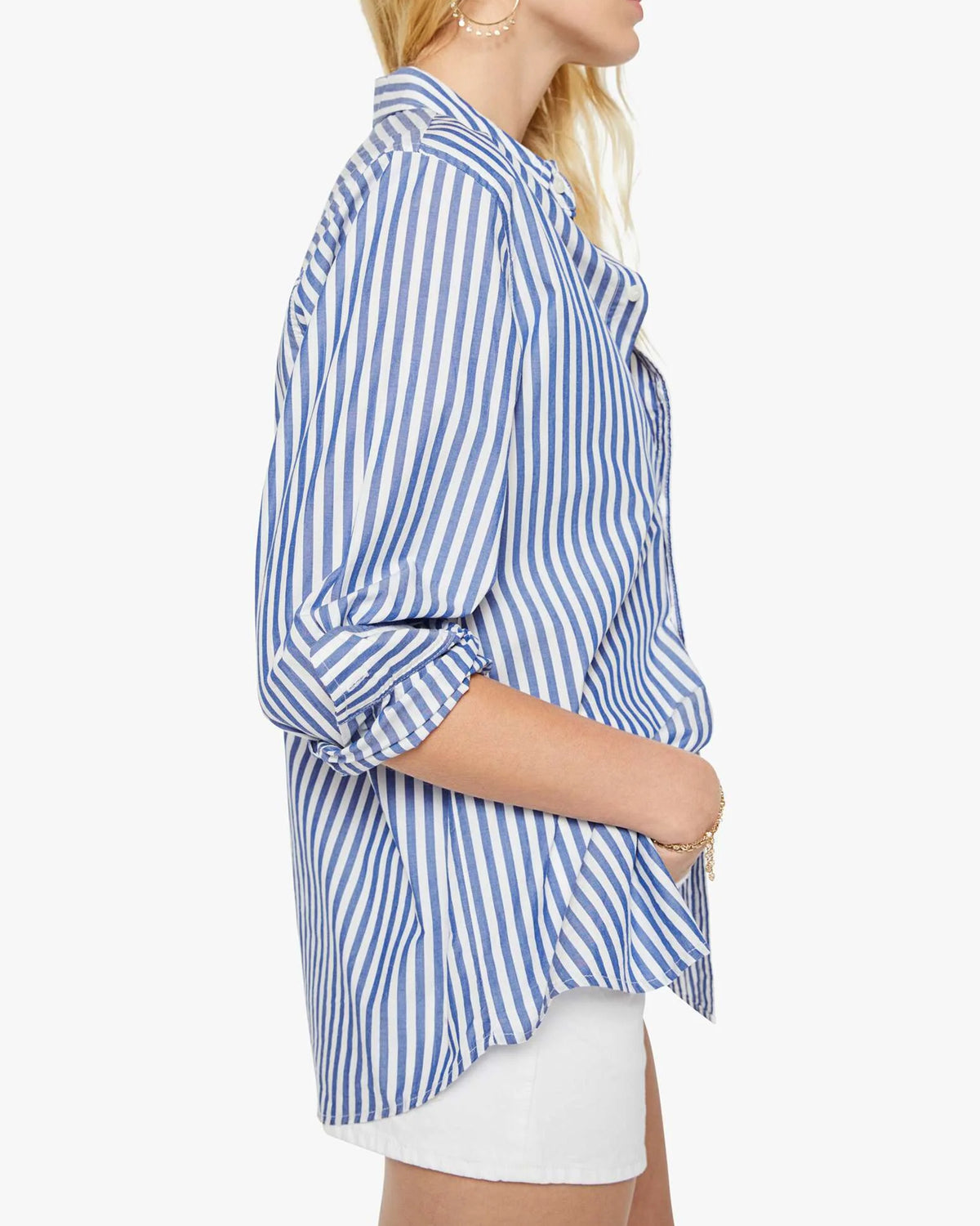 A woman poses in profile against a white background, wearing The Bestie blue and white striped cotton button-up shirt with rolled sleeves from Mother, paired with white shorts.