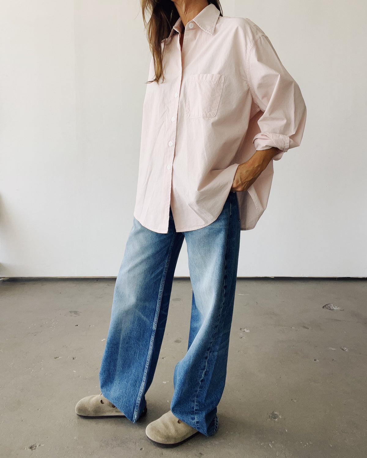 Wearing Hey Gang's The Big Shirt in light pink, paired with baggy blue jeans and beige slip-ons, the person stands indoors against a minimal background, showcasing a relaxed, made-in-Los Angeles vibe.