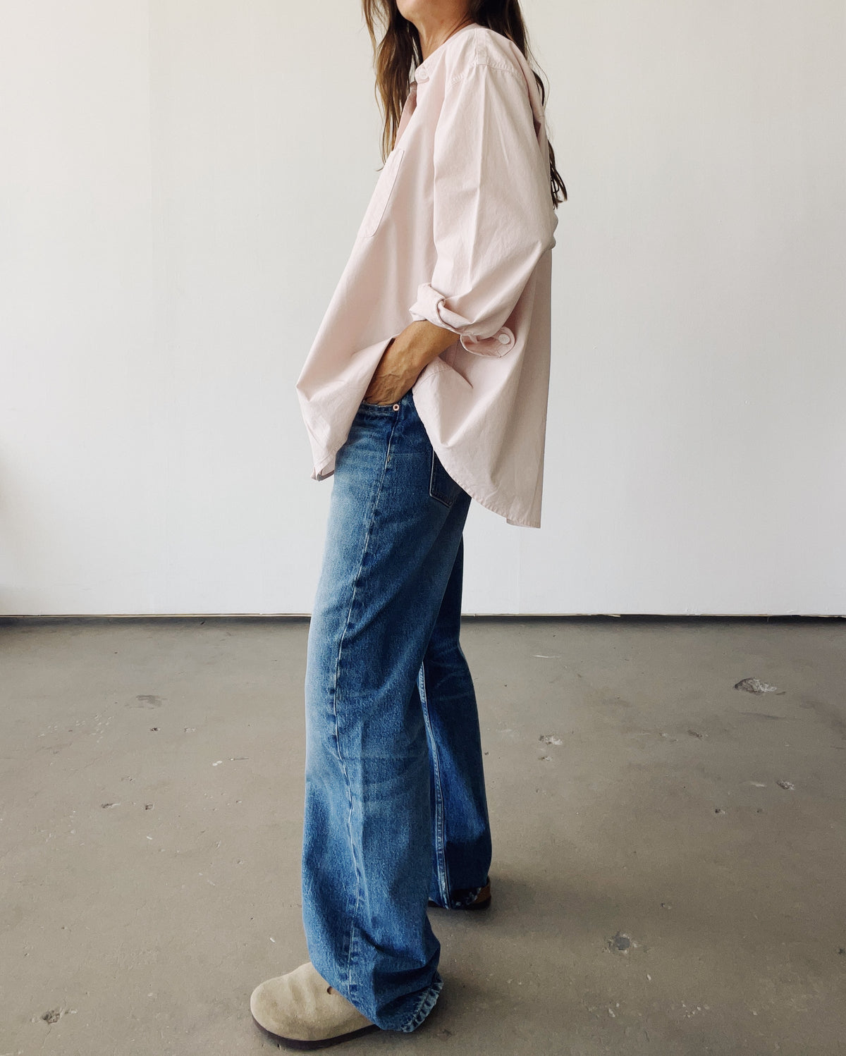 A person stands indoors against a plain white wall, wearing Hey Gang's oversized The Big Shirt in light pink cotton poplin with rolled-up sleeves, blue wide-leg jeans, and beige shoes. Their hand is in a pocket and their face is partially out of view.