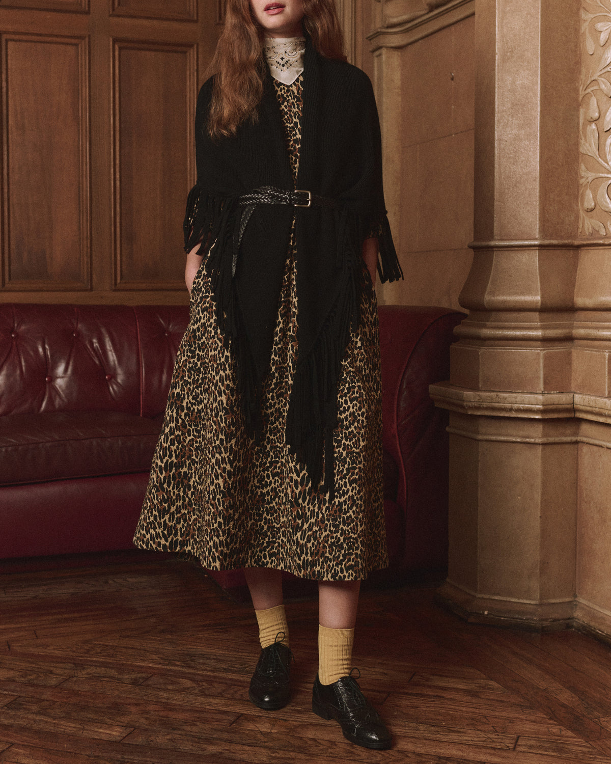 Leopard print dress with The Great Inc. Big Sweater Fringe Scarf, styled in an elegant, ornate room by a red couch.