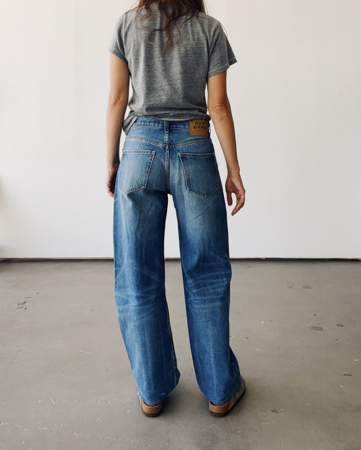 A woman stands indoors facing away, wearing Hey Gang's The Wide Slouch Jean in Harris wash with a gray t-shirt and brown shoes, set against a plain white wall and concrete floor.