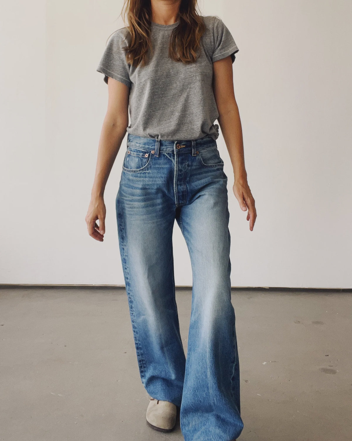 A person stands indoors against a plain light wall, wearing the Hey Gang Wide Slouch Jean in a loose fit with a gray t-shirt and beige shoes. Their face is partially out of frame.