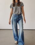 A person stands indoors against a plain light wall, wearing the Hey Gang Wide Slouch Jean in a loose fit with a gray t-shirt and beige shoes. Their face is partially out of frame.